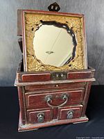 Front view of the antique Chinese jewelry box with hidden mirror lid opened, showing carved floral gold leaf frame around mirror and multiple drawers with metal handles.