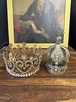 Two crowns displayed side by side against a framed painting backdrop