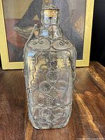 Front view of the antique metal bound filigree worked bottle on a wooden surface with a framed painting in the background.