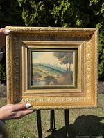 Full front view of the oil painting on metal with gilt frame held outdoors