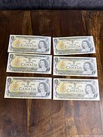 Front side of six uncirculated Canadian $1 bank notes arranged in pairs on a wooden surface, showing Queen Elizabeth II portrait and detailed design.