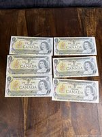 Six Canadian one dollar bills displayed face up showing Queen Elizabeth II and Canadian coat of arms.