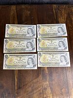 Front sides of six Canadian $1 banknotes laid in two rows of three on wooden surface showing Queen Elizabeth II and coat of arms.