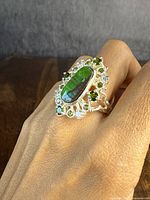 Front view of the sterling silver ring worn on hand showing the central elongated oval Ammolite with surrounding green garnets and clear stones in cluster setting.