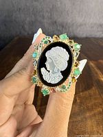 Front and side views of the sterling silver ring, highlighting the detailed carved mother of pearl cameo of a woman's profile on a black base and decorated with green and pink natural colored gemstones.