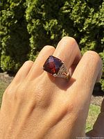 Ring worn on finger in natural light showing large square-cut garnet center stone and clear stones on sides with gold overlay on sterling silver band.