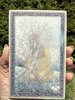 Close-up of plaque held in hand showing detailed painting of seated man in traditional Mughal attire with floral background.