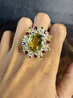 Ring on finger showing large central yellow gemstone with surrounding red, green, and light yellow stones.
