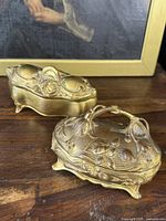 Pair of two ornate antique Art Nouveau style metal dresser jewelry boxes on wooden surface with framed artwork in background, front view showing detailed floral motifs and lid shapes.
