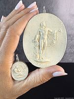 Hand holding two oval carved plaster cameo pendants showing entire works and size comparison.
