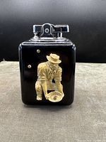 Front view of the black Bakelite Ronson table lighter showing the beige-colored 3D relief of a crouching gold miner panning for gold.