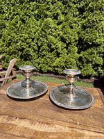 Pair of silver-plated chamber candlestick holders displayed outdoors on a wooden surface with green foliage background.