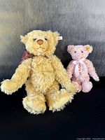 Two Steiff teddy bears standing against a dark backdrop, larger yellow bear on left and smaller pink bear on right.