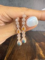 Close-up photo of the pair of earrings with circular clear stones, pear-shaped orange stones, and natural opal drops in rose-gold overlay, held between fingers.