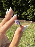 Ring held between fingers showing the purple stone and side clear stones with ornate band design, outdoors in daylight.