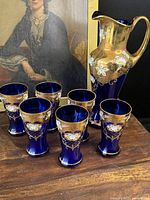 Photo showing the full set with the tall pitcher and six glasses arranged on a wooden surface, a painting in the background.