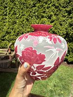 Image showing bulbous cameo glass vase held by hand outdoors against greenery, highlighting red floral and leaf motifs on frosted translucent background with flared neck.