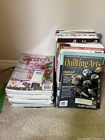 Two stacks of magazines on floor; left stack showing Stitch magazine at top, right stack showing Quilting Arts magazine on top