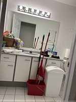 Wide view of bathroom sink area with cleaning tools: red mop bucket with mop and broom, white trash can with lid, visible countertop with supplies.