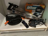 BLACK+DECKER Mouse MSS500K electric sander with box, black molded plastic storage case with orange latch, a metal hand saw with a black grip handle, and a rectangular bubble level.