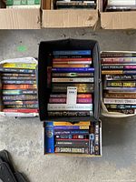 Top-down view of three boxes/bins with books arranged closely together showing spines and covers of various fiction and thriller titles.