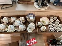Wide view of lot including teacup and saucer sets, several plates, mugs, and cream and sugar dishes arranged in cardboard boxes on a kitchen counter.