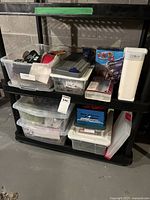 Plastic storage bins on black plastic shelving with a variety of crafting items including ribbons, a box labeled 'Tracer Projector Jr.', and an OttLite rechargeable light.