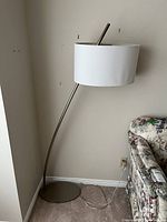 Full view of modern curved metal floor lamp with off-white fabric drum shade standing near a wall and a sofa.