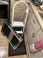 Photo of the foldable cart with tape repairs on the handle, white step stool, and black floor mat placed on kitchen floor.