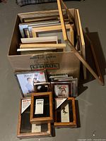 Box filled with numerous picture frames of various sizes and styles, including wood and metal finishes. Three new instant shadow box frames with wood finish displayed in front of the box.