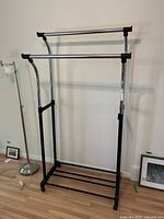 Frontal view of the metal garment rack showing two horizontal hanging bars and black metal base on wooden floor with wall background.