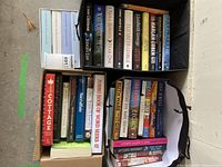 Boxes and bags containing a mixed collection of hardcover and paperback books by various notable authors. The books cover multiple genres and show some signs of light use.