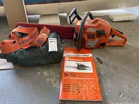 Full lot arranged on concrete floor showing chainsaw, bar scabbard, safety manual and chaps bag with straps
