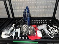 Photo showing the blue T-fal iron, transparent Clover craft iron, white hair dryer, and small black accessory item with cords on a black metal rack.