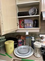 Kitchen corner showing assortment of kitchen items including decorative tins in upper cabinet, glassware, plates, pitchers, yellow waste bin, and containers on counter
