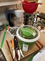 Wide photo showing Black & Decker can opener, red colander, salad spinner, cutting boards, spice rack, knives, and other kitchen utensils on countertop.