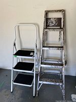 View of both ladders side by side against a wall, showing the heavily stained metal ladder and the white step stool ladder.