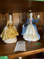 Two Royal Doulton bone china figurines, Coralie in yellow dress on left and Leading Lady in blue and white dress on right, displayed on wood shelf.