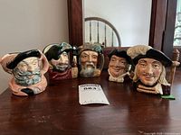 Five Royal Doulton Toby Jugs displayed on a wooden table, showing various characters including Falstaff and Robinson Crusoe.