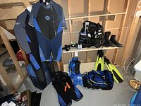 Two wetsuits hanging on hangers showing front and back views, dive boots on shelf along with some diving accessories, fins and gear bag on floor.