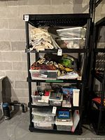 Front view of black plastic shelving unit filled with various plastic containers and folded fabrics; shelf sold without contents.