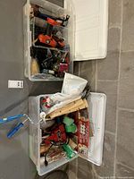 Lot of assorted tools and painting supplies packed in transparent plastic containers, showing power drill, hammers, tape rolls, paintbrushes, and other items
