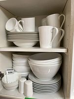 White dinnerware set placed on shelving including stacked plates, bowls, mugs, and salt and pepper shakers.