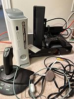 Xbox One and Xbox 360 consoles with headphone and joystick controller placed on floor with cables.