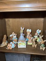 Wooden shelf displaying a collection of 11 bunny figurines of various sizes and materials, showing upright, sitting, and laying poses with pastel color clothing.