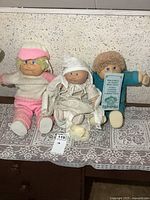 Three Cabbage Patch dolls sitting on a lace cloth against a wall, showing a range of outfits and hair colors.