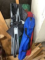 Four folded camp chairs with metal frames and fabric seats packed in black, green, red, and blue carrying bags leaning against a wall.