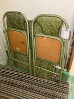 Rear view of two folding chairs stacked, showing green vinyl upholstery with mottled pattern and metal tubular frames