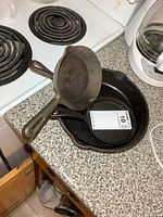 Three cast iron pans stacked on kitchen counter near stove. Largest pan is Smart Brockville 9-inch with visible wear.