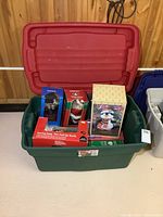 Green Rubbermaid Rough Tote with red lid, filled with boxed animated Christmas figures like Santa in fire truck, penguin, and others.